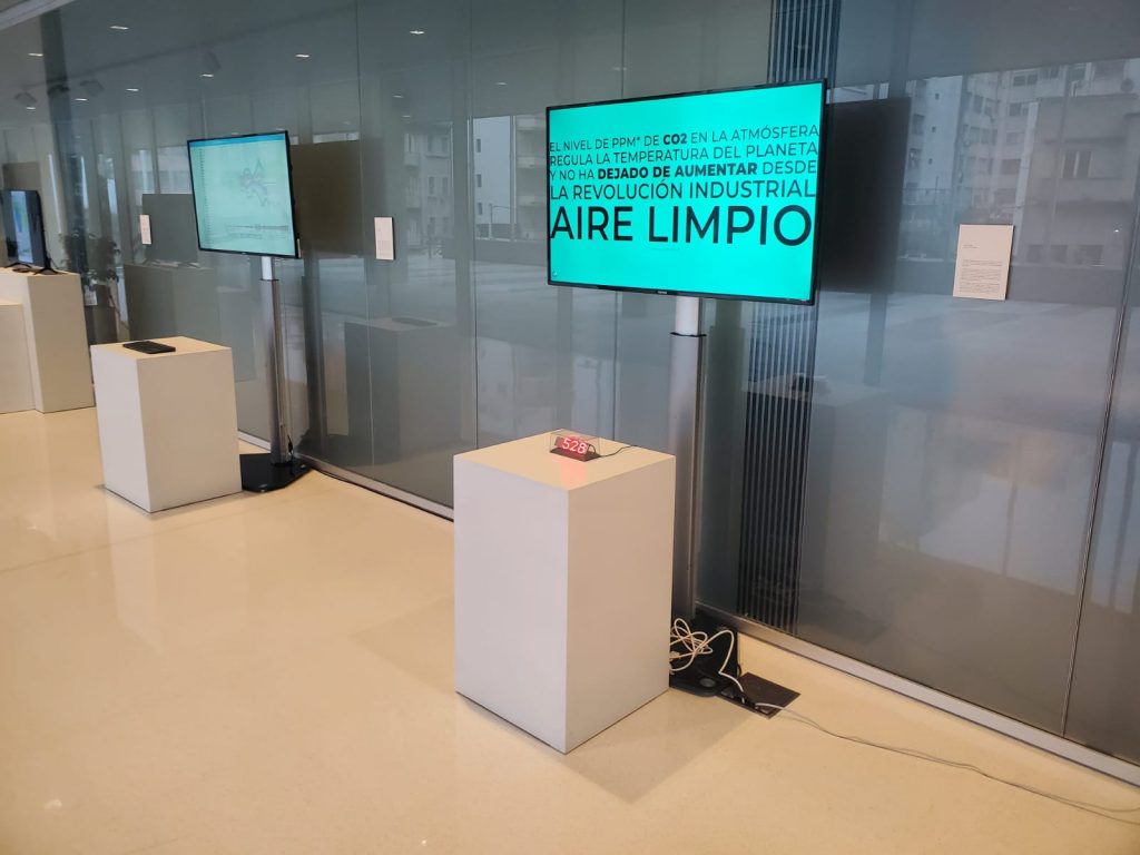 Instalación de la obra, con un pedestal que sostiene un display LED indicando la cantidad de CO2 en el aire y una pantalla con información detrás.